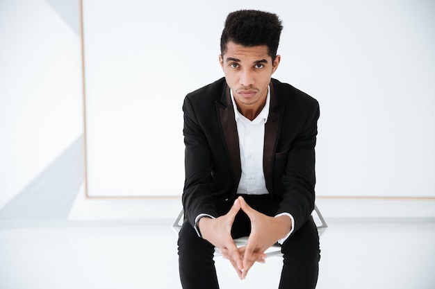 Serious African business man in black suit sitting on chair in office and looking at camera