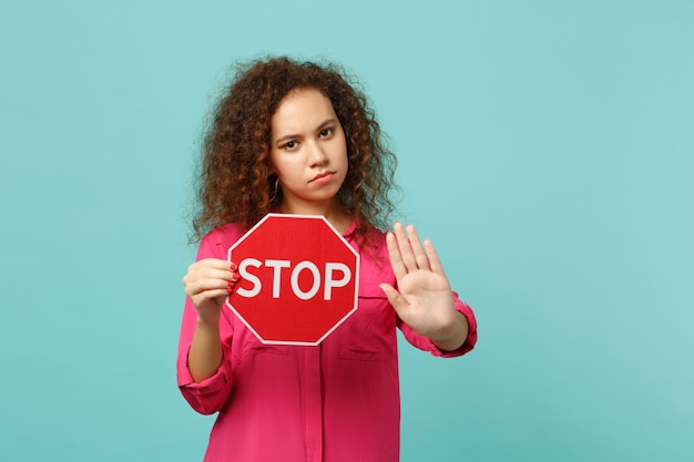 파란색 청록색 배경에 격리된 손바닥으로 정지 제스처를 보여주는 STOP 텍스트 보드를 들고 분홍색 캐주얼 옷을 입은 진지한 아프리카 소녀. 사람들은 진심 어린 감정 라이프 스타일 개념입니다. 복사 공간을 비웃습니다.