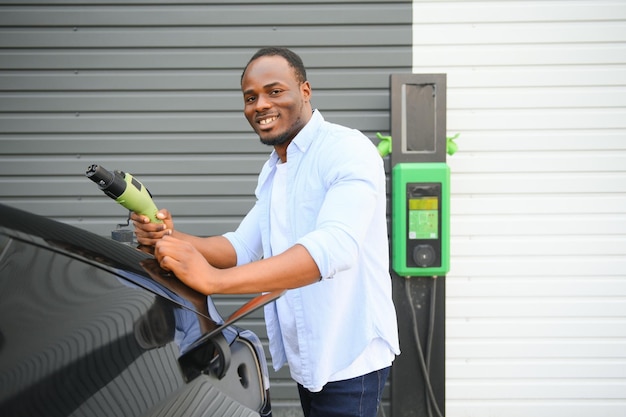 高級電気自動車の近くに立って、充電ケーブルを手に持った深刻なアフリカ人男性