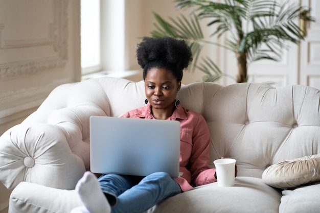 Serious african woman work on laptop sit on sofa with coffee or tea relaxing after remote occupation