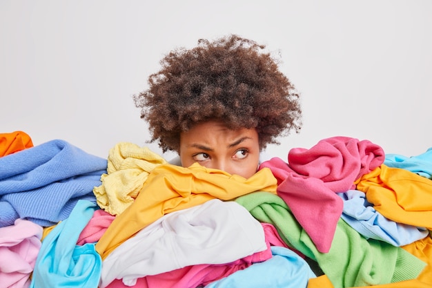 Photo serious afro american woman poses near heap of multicolored unsorted clothes after washing brings order in closet looks attentively away isolated over white wall collects clothing for sale