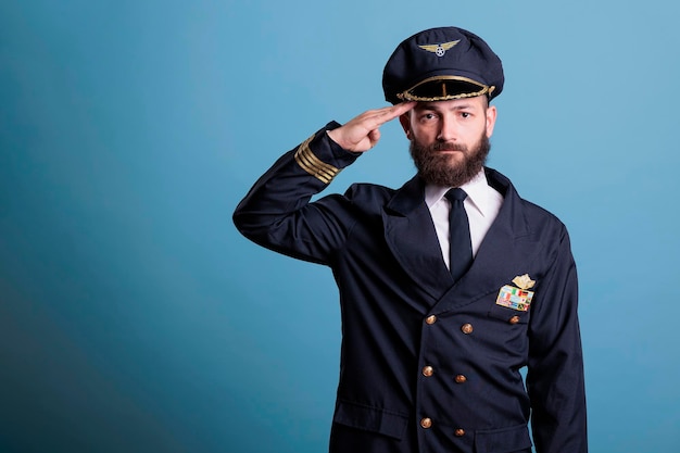 Serious airplane pilot saluting, wearing uniform and hat front view portrait, plane captain looking at camera. Aviation academy aviator with airline wings badge on jacket