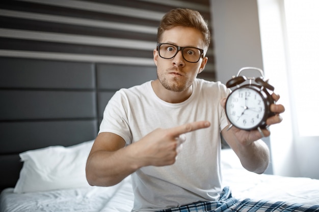 Foto giovane arrabbiato serio sul letto nella mattina. guarda la telecamera e indica l'orologio.