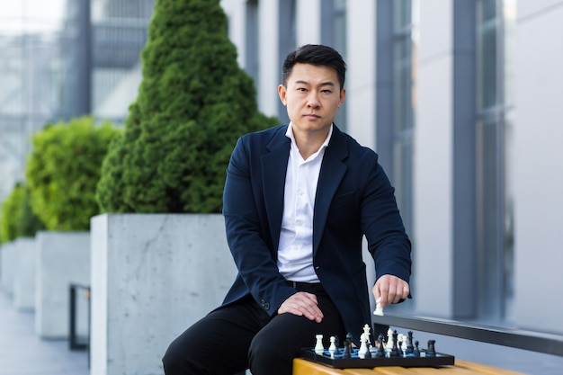 Serious asian man playing chess outside, businessman thinking playing chess sitting on bench near office center