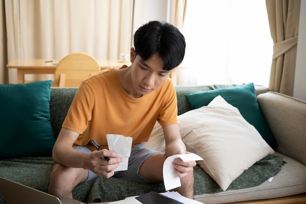 Серьезный азиат сидит на диване и подсчитывает домашние расходы