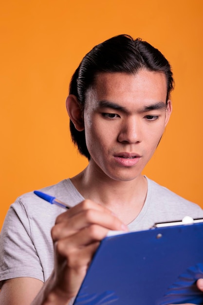 파란색 클립보드에 메모를 하는 진지한 아시아 남자, 메모장에 쓰는 똑똑한 학생이 닫힙니다. 설문지를 작성하는 집중된 젊은이, 문서를 들고 있는 회사 직원