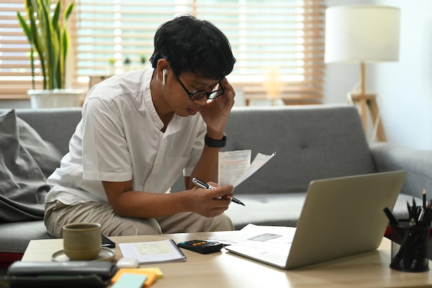 Serious asian man using laptop computer and calculating household expenses