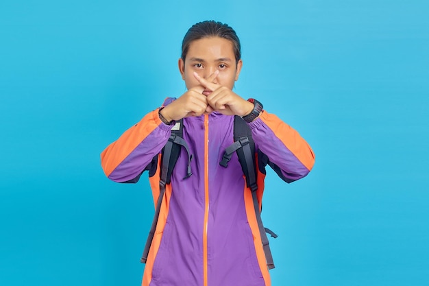 Serious asian young man showing refusal gesture with crossed fingers isolated on blue background