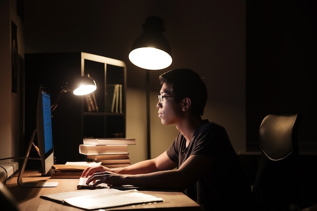 Serious asian young man using computer and typing in dark room