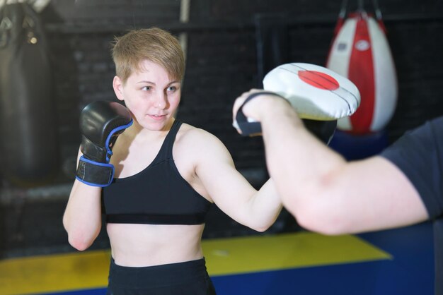 真面目なアスレチックスポーティーフィットフィットネス強い美しい女の子の戦闘機は、手袋で戦うボックストレーニングを持っているジムで若い女性のトレーニングをボクシングしていますトレーナーとスパーリング健康的なスポーツライフスタイル