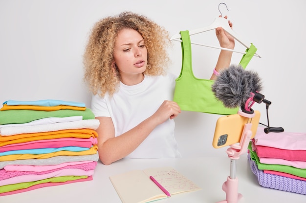 Serious attentive curly haired female entrepreneur sells clothes online advertises green top on hangers poses at table with colorful folded laundry isolated over white wall. Female influencer