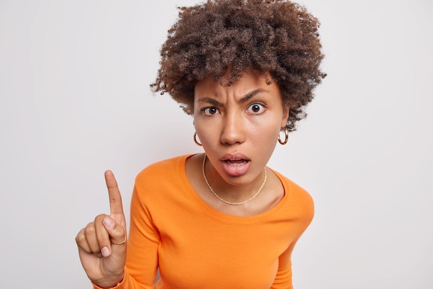 Serious attentive curly haired woman has idea points index finger above shows direction wears casual orange jumper and earrings poses against white studio background You should check this out