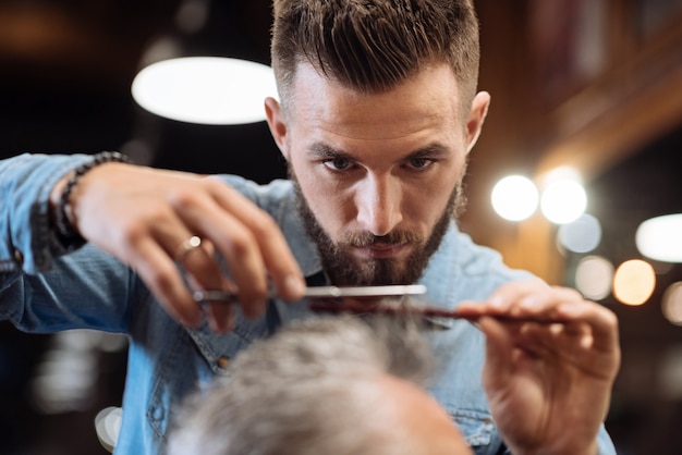 Foto atteggiamento serio. ritratto del primo piano di bello giovane parrucchiere barbuto che tiene forbici e pettine mentre taglia i capelli del suo cliente.