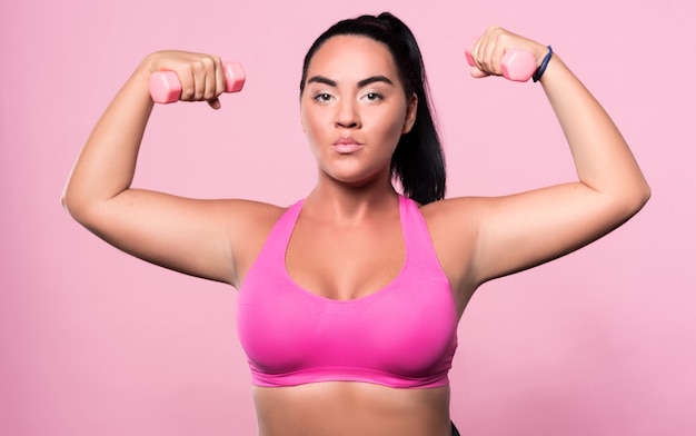 Serious attitude. Portrait of brave pretty mulatto woman standing with small dumbbells in both her hands isolated