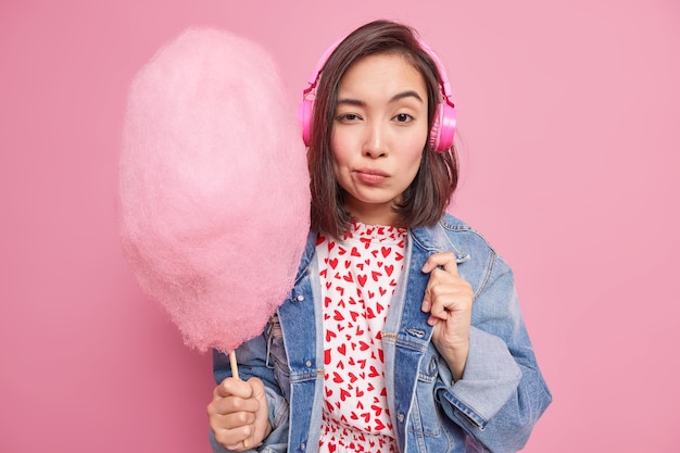 Serious attractive Asian woman looks confidently  holds candy floss listens music via headphones dressed in stylish clothes isolated over pink wall. Teenage lifestyle concept.