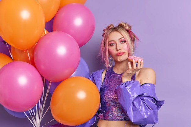 Serious attractive young European woman looks bored, dressed in fashionable purple jacket and cropped top holds tasty donut being on party poses with bunch of colorful helium balloons