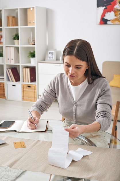 Foto giovane donna attraente seria che si siede al tavolo e che visualizza il controllo finanziario mentre registra le spese nel diario