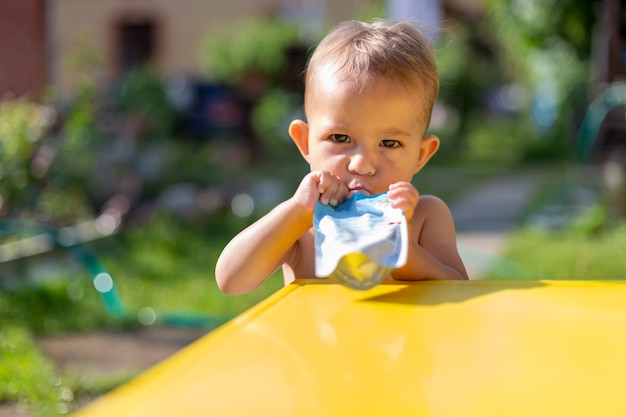 ポーチでフルーツピューレを食べる深刻な赤ちゃんは晴れた日にカメラの黄色いテーブル緑の庭をのぞきます