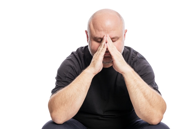 A serious bald middle-aged man in a black T-shirt is sitting with his hand in his face. Isolated over white .