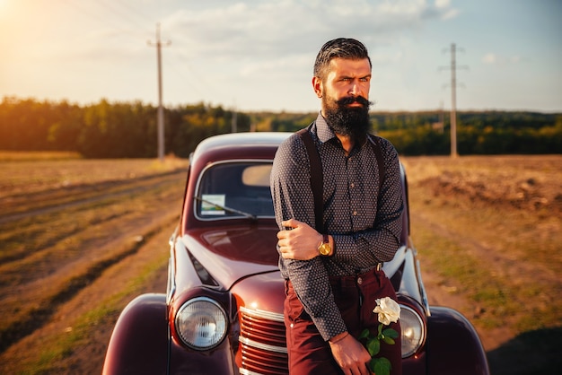 Serious bearded brunette man in a shirt, trousers with suspenders near a brown retro car on the background of a country road and sunset