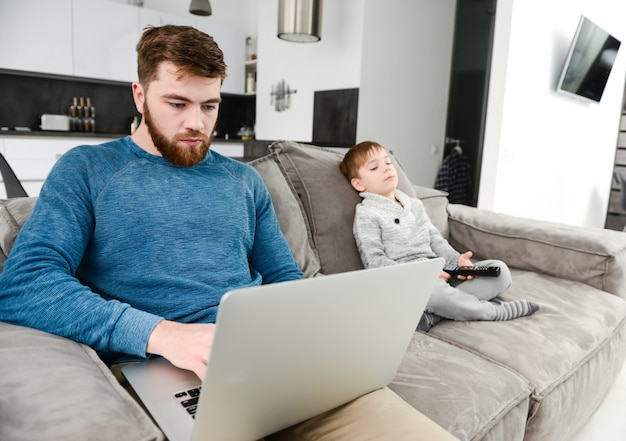 Padre barbuto serio vestito con un maglione blu usando il portatile mentre suo figlio guarda la tv.