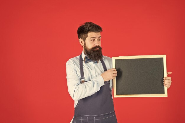 Serious bearded man in barber apron hold barbershop board red background copy space school