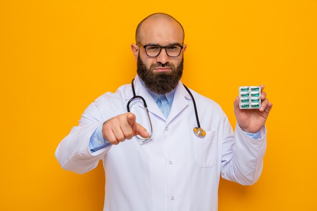 Grave uomo barbuto medico in camice bianco con stetoscopio intorno al collo con gli occhiali che tengono blister con pillole puntate con il dito indice