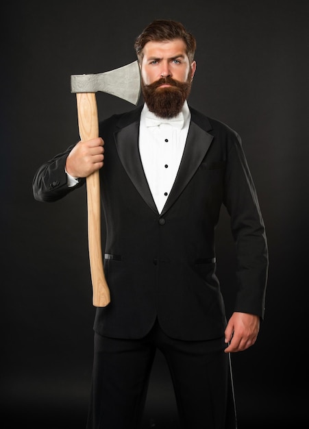 Serious bearded man in formal suit holding axe at unshaven face dark background barber