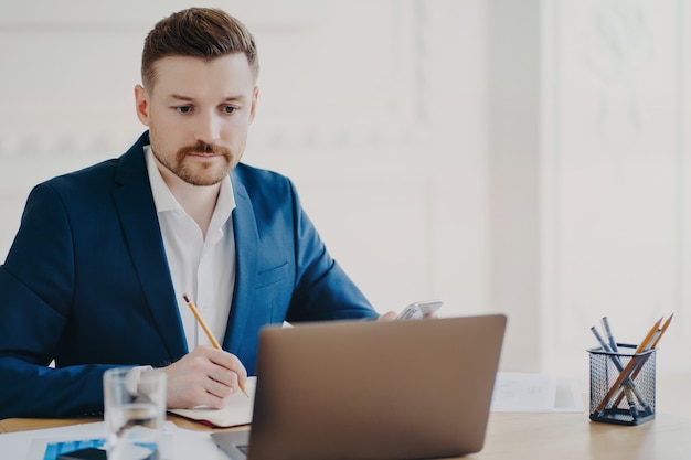 Serious bearded man marketer involved in working process makes notes with pencil looks attentively at laptop computer wears blue formal suit poses at coworking space writes organisation plan