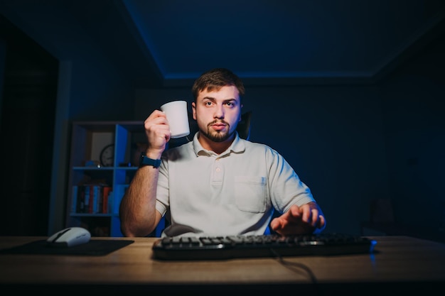 Serious bearded man online worker working at night on the
computer at home with a cup of drink