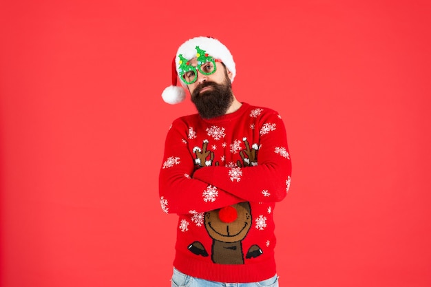 Serious bearded man in santa claus hat and party glasses on red background, winter holiday