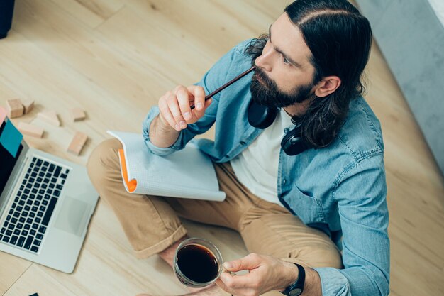 Serious bearded man sitting in front of a laptop with a cup of coffee and touching his nose with a tip of a pencil