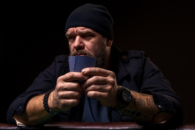 Serious bearded man sitting at poker table and holding cards isolated on black