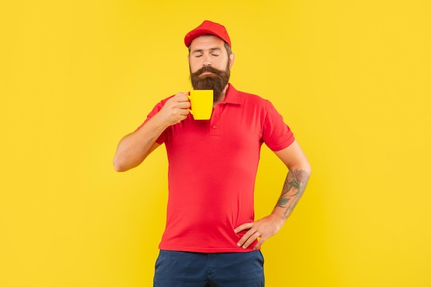 Serious bearded man smelling hot beverage with closed eyes holding cup yellow background coffee