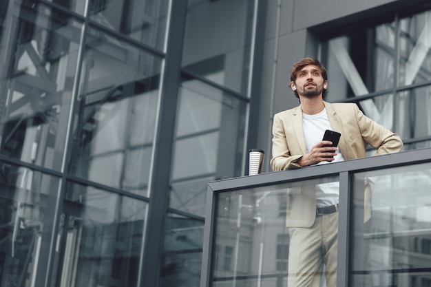 Serious bearded man in stylish suit and earphones holding smartphone and looking straight while standing outdoors