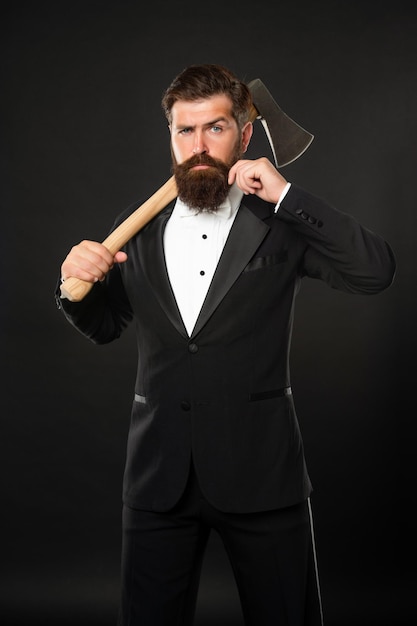 Serious bearded man twirling mustache while carrying axe at dark background barber