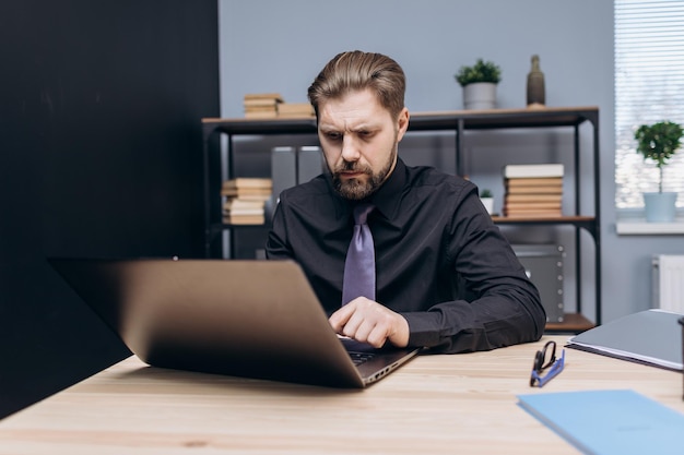 Серьезный бородатый мужчина работает на ноутбуке в офисе