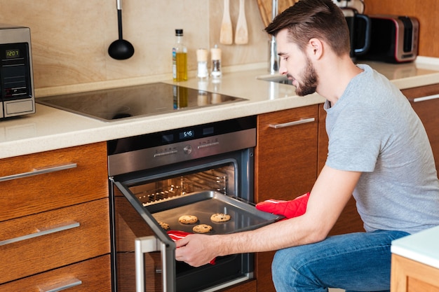 Photo serious bearded young man baking cookies in oven on the kitchen