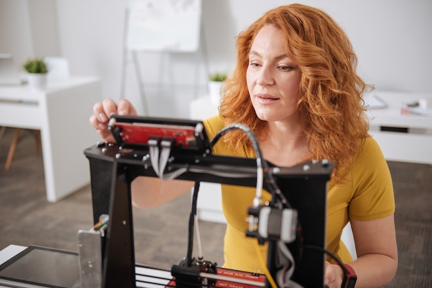 Foto grave bella donna concentrata seduta davanti alla stampante 3d e la configurazione mentre si concentra sul suo compito