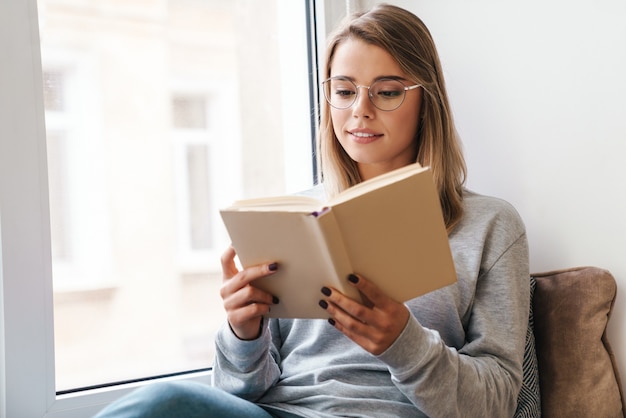 serious beautiful woman in eyeglasses reading book while sitting near window indoor