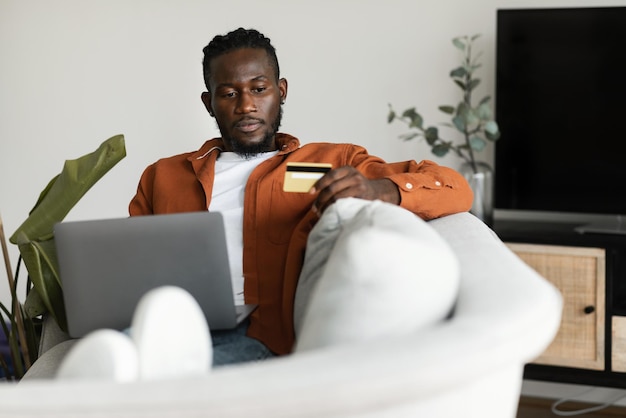 Uomo di colore serio che utilizza la carta di credito per lo shopping online seduto sul divano con lo spazio libero interno della casa del computer portatile