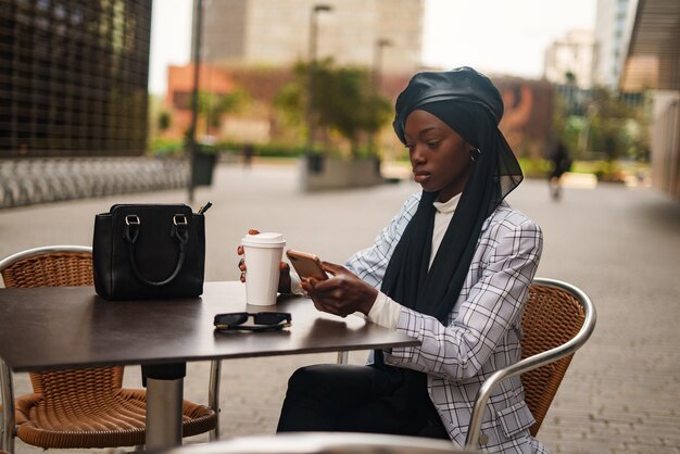 Serious black woman using smartphone at table in outdoor\
cafe