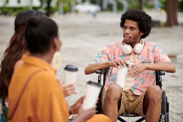 Серьезный черный молодой человек с инвалидностью пьет кофе и слушает историю своего друга