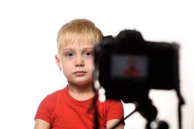 Serious blond boy is recording a video on DSLR camera. Little video blogger. White background