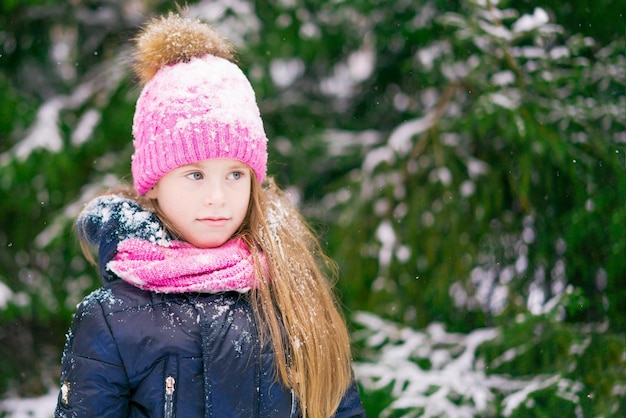 A serious blond girl kid in pink hat in a winter forest