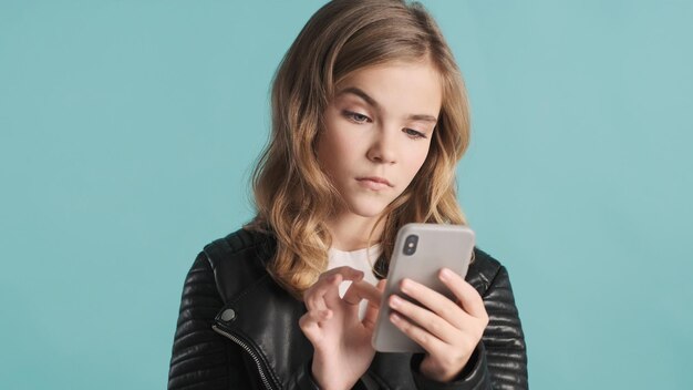 Serious blond teenage girl raising brow chatting on smartphone over blue background. Dissatisfied face expression