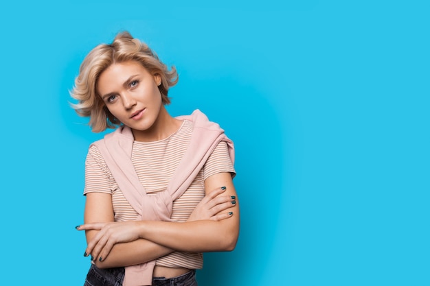 Serious blonde woman with curly hair posing with crossed hands on a blue wall