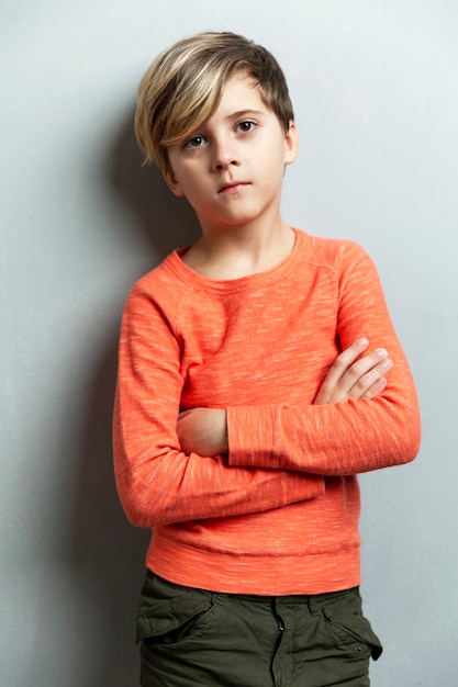 A serious boy of 9 years old in an orange jacket stands against the wall. The arms are crossed over the chest. Gray background. Vertical.