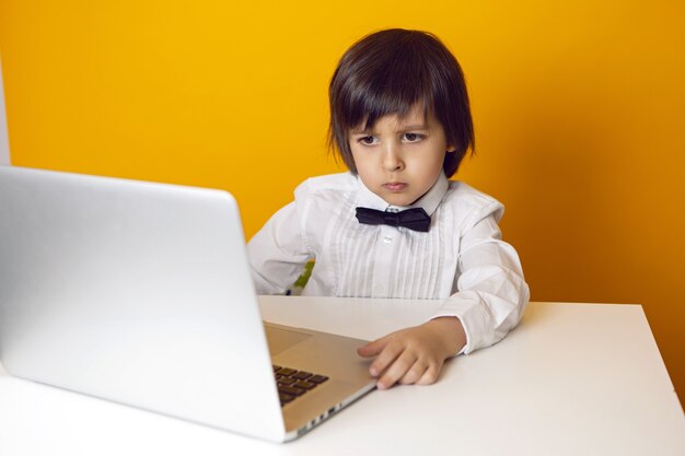 Serious boy child businessman in a white shirt and bow tie sits at a laptop at a desk in a yellow office