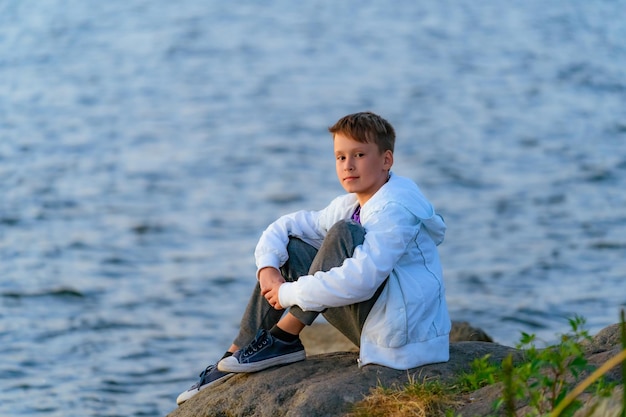 후드와 청바지를 입은 흰색 재킷을 입은 진지한 소년이 강 근처의 바위에 앉아 있습니다. 확대.